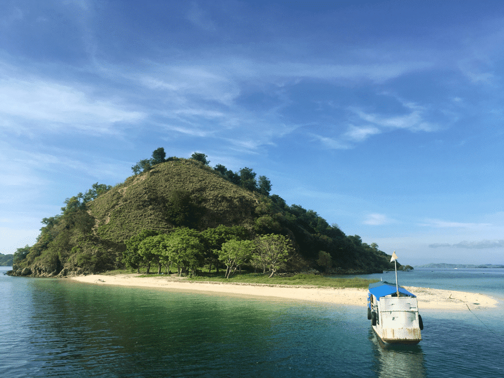 Kelor Island. Scenic island with green hill, sandy beach, and anchored boat under a clear blue sky. Peaceful tropical landscape.