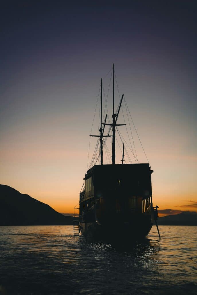 komodo island trekking. Silhouette of a sailing ship at sunset on the ocean with a serene orange sky background and distant mountains.