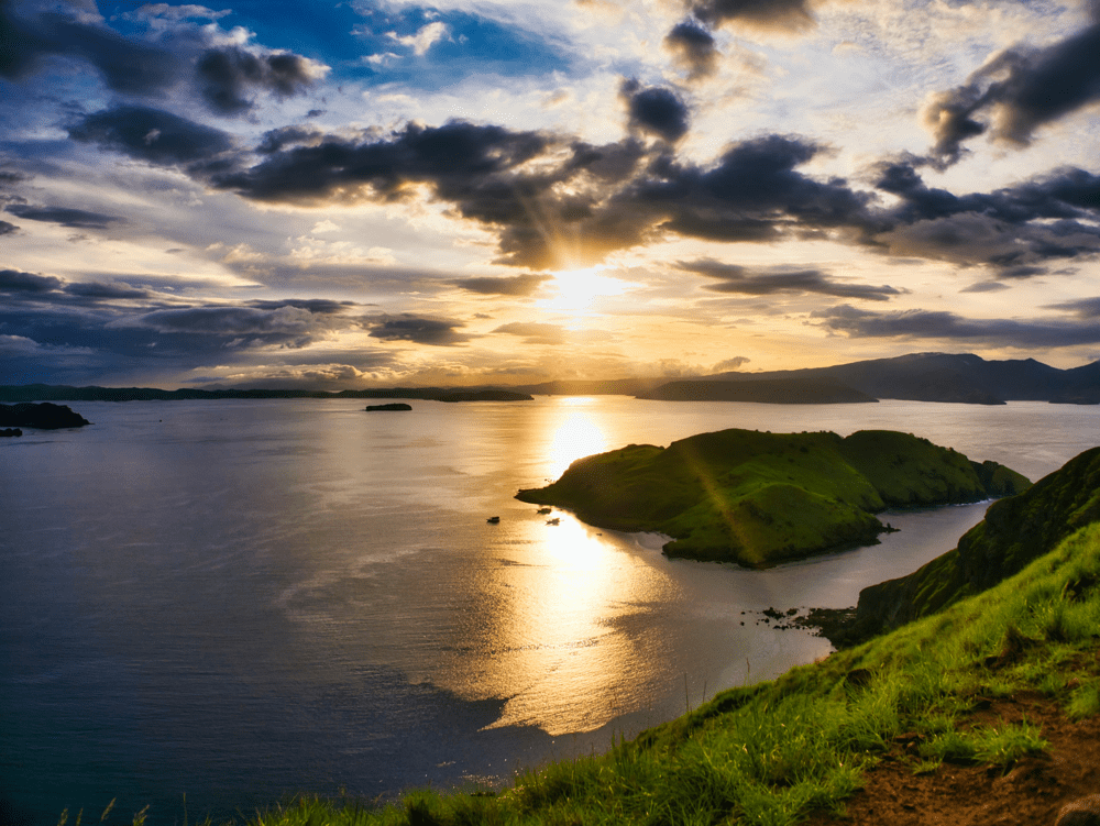 Padar Island Indonesia – Sunset over a serene bay with lush green hills, clouds reflecting golden light on calm waters.