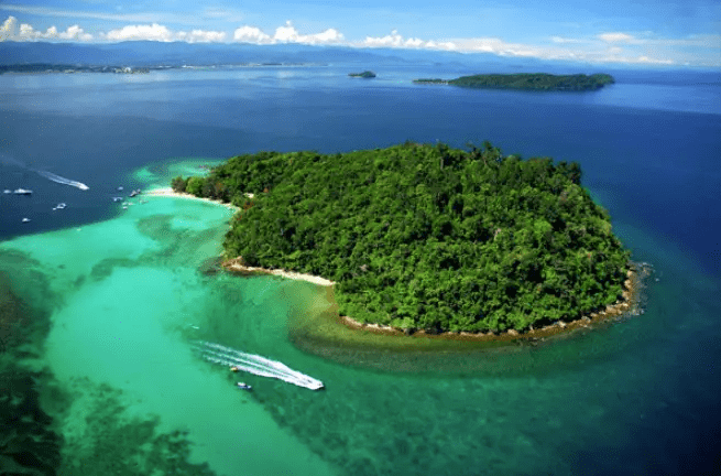 Sabolo Island. Aerial view of a lush green island surrounded by clear turquoise waters and small boats in the distance.