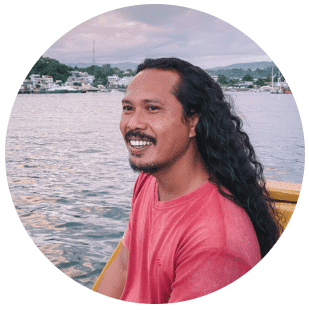 Yanto - Indonesianboattrip.com -A smiling man with long hair sits on a boat, overlooking a calm waterway and a scenic shoreline at sunset.